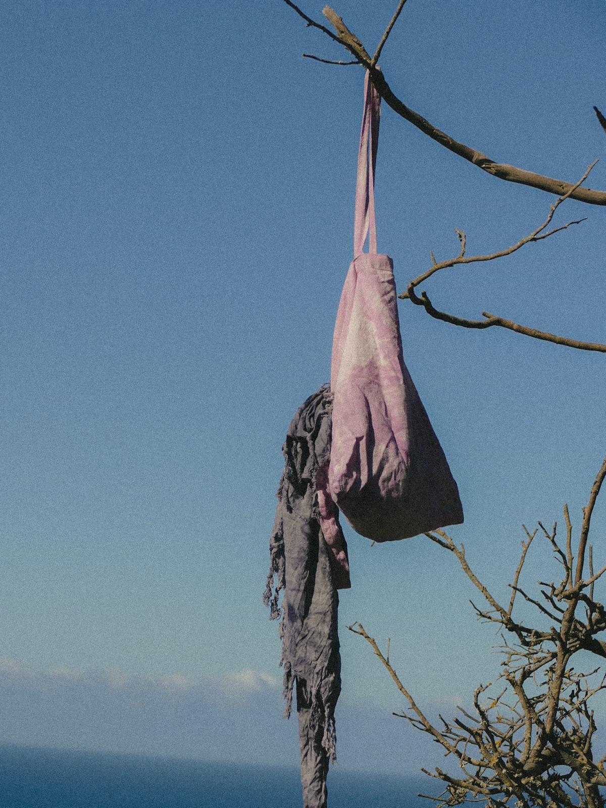 Lilac Cocou tote bag and grey sarong hanging from a tree branch with an ocean backdrop. Hand dyed Jute bag Lilac