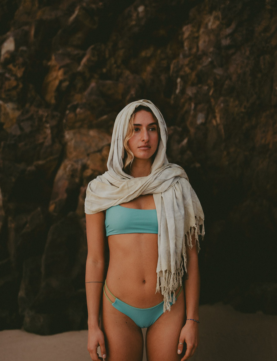 Woman in a light blue bikini with a natural fabric shawl draped over her head, standing against a rocky background on the beach.