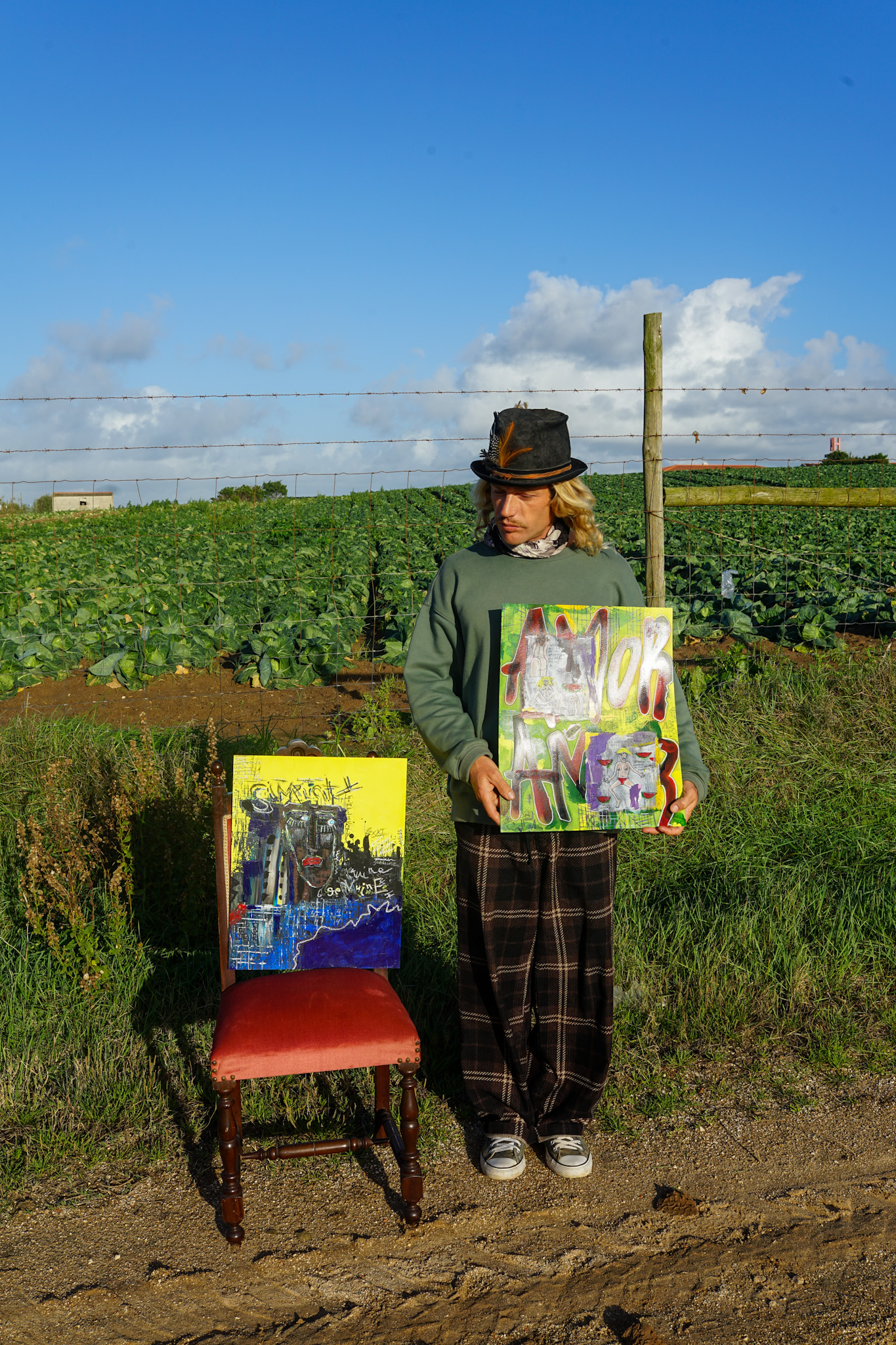 Rebelglimpse standing outdoors in front of a lush green field, holding a colorful abstract painting, with another piece of his artwork displayed on a chair beside him.