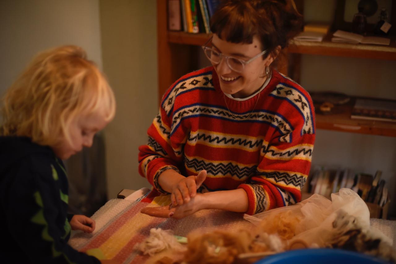Theresa, wearing a colorful sweater, smiles as she works on a felt piece with a young child, surrounded by wool and tools.