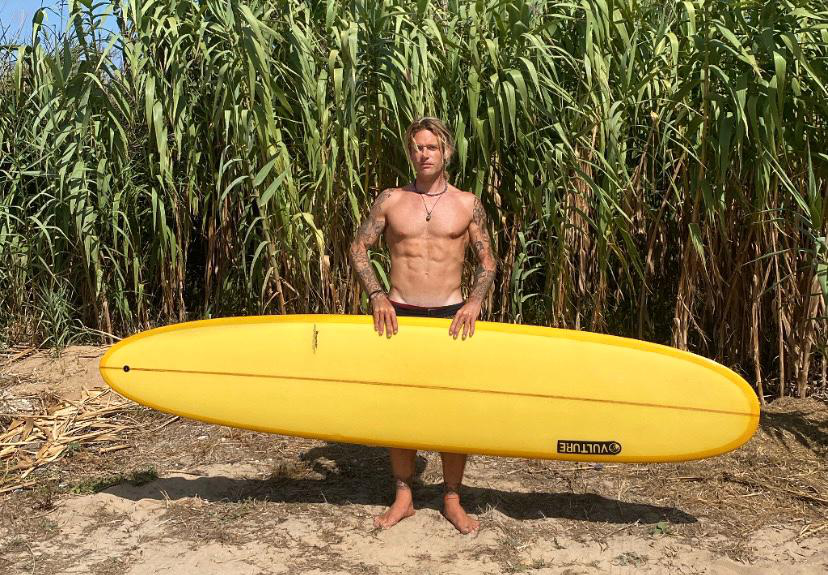 Rebelglimpse holding a vibrant yellow longboard, standing barefoot on a sandy path against a backdrop of tall green reeds under a bright blue sky.