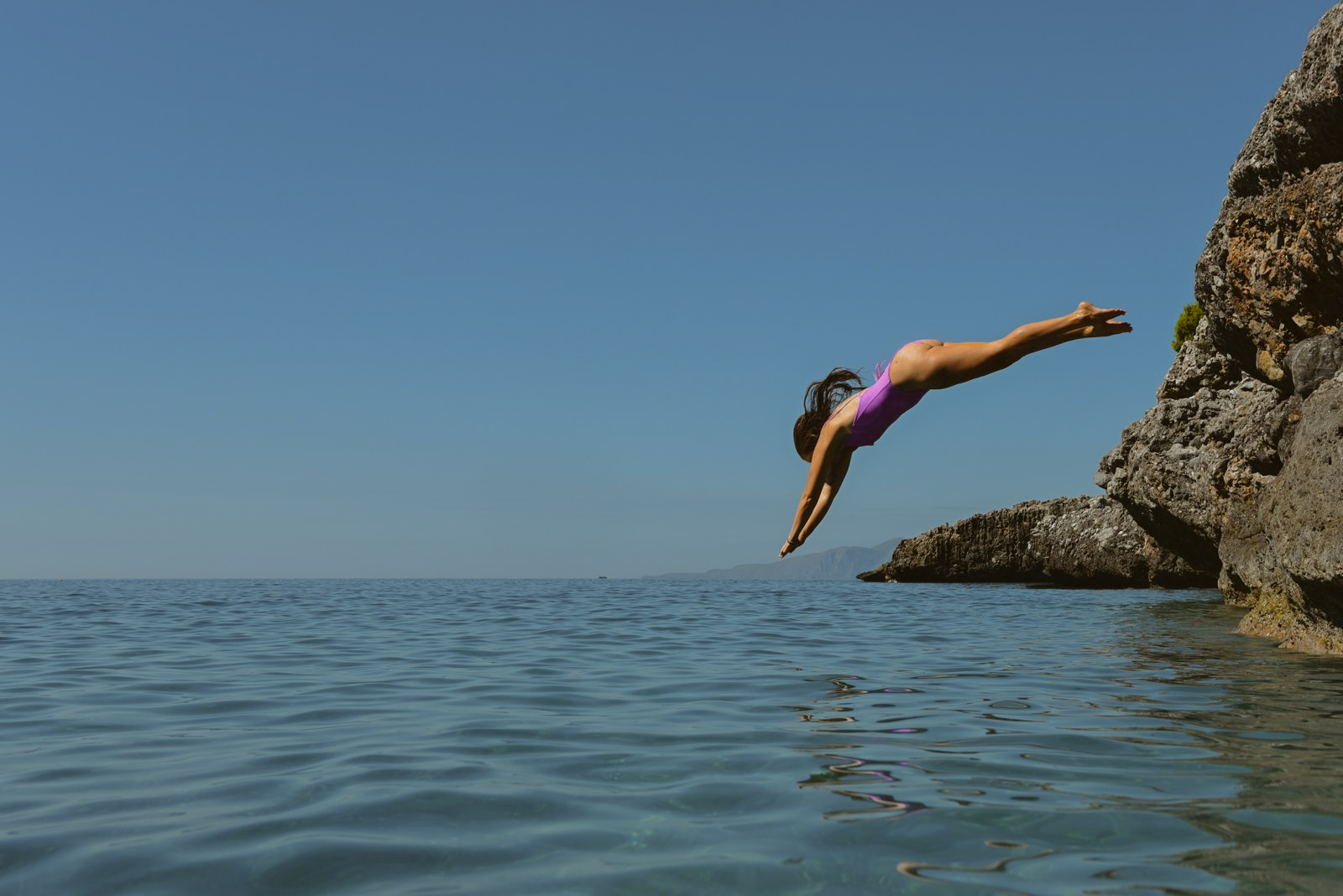 Female founder of Sustainable Swimwear in a reversible lilac bikini diving off a rocky cliff into the ocean, representing freedom, adventure, and sustainable swimwear. The bikini is ethically handmade in Italy from recycled materials, embodying slow fashion and eco-conscious design. [Sustainable swimwear], [ethical bikinis], [reversible bikini], [eco-friendly fashion], [slow fashion], [female-owned brand], [handmade in Italy], [conscious couture], [minimal swimwear], [adjustable bikini]