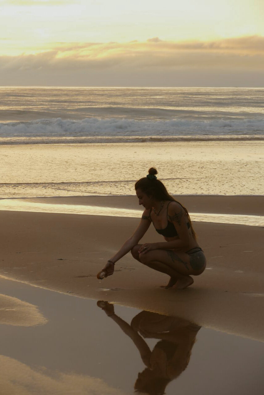 Mindful Beach Moments – Ethical Swimwear for Ocean Lovers | Cocou Conscious Couture A female founder of a sustainable swimwear crouching on the beach at sunset, picking up a seashell. She wears a black sustainable bikini from Cocou Conscious Couture, reflecting an eco-conscious and minimalist lifestyle. Her reflection in the wet sand symbolizes a deep connection with nature and mindful living.